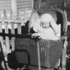 A baby boy in a wicker baby carriage. The baby is wearing light color clothing including a hat, long sleeves, and gloves. The baby is in bright sunlight and his eyes are nearly closed.