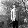 A young man, probably 16 years old, standing outside in front of some trees and bushes with houses in the background. He is wearing a sports coat, sweater vest, and tie.
