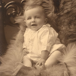 A smiling baby sits on a chair facing the camera. The baby is sitting on a thick blanket or other woven material, possibly a fur throw.