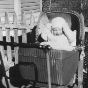 A baby boy in a wicker baby carriage. The baby is wearing light color clothing including a hat, long sleeves, and gloves. The baby is in bright sunlight and his eyes are nearly closed.