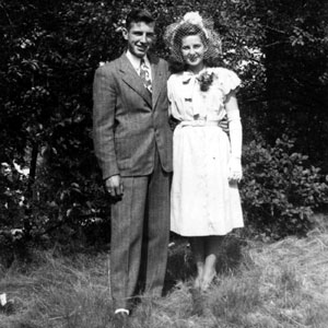 A young couple standing arm-in-arm in front of trees and bushes. The man is on the left in a suit and tie. The woman is on the left wearing a white dress, long white gloves, and a corsage.